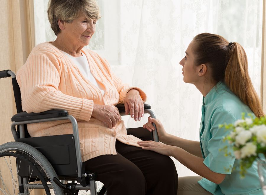 A caregiver kneeling beside an older woman, holding her hand and listening